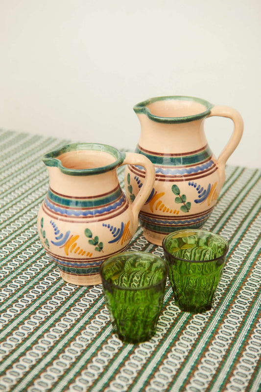 Dos jarras de cerámica artesanal con motivos florales y dos vasos de vidrio verde sobre un mantel a rayas.