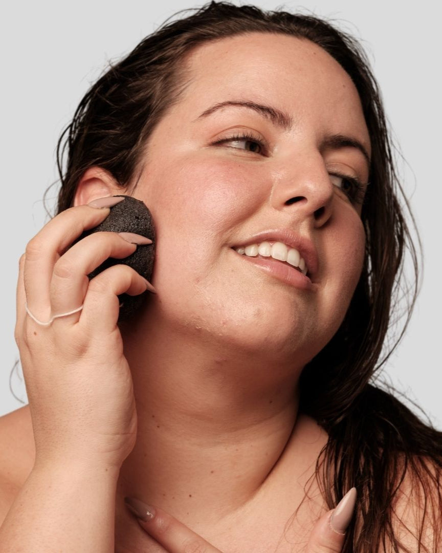 Mujer usando una esponja facial Konjac negra de Banbu para limpiar su rostro con suavidad sobre fondo gris neutro.