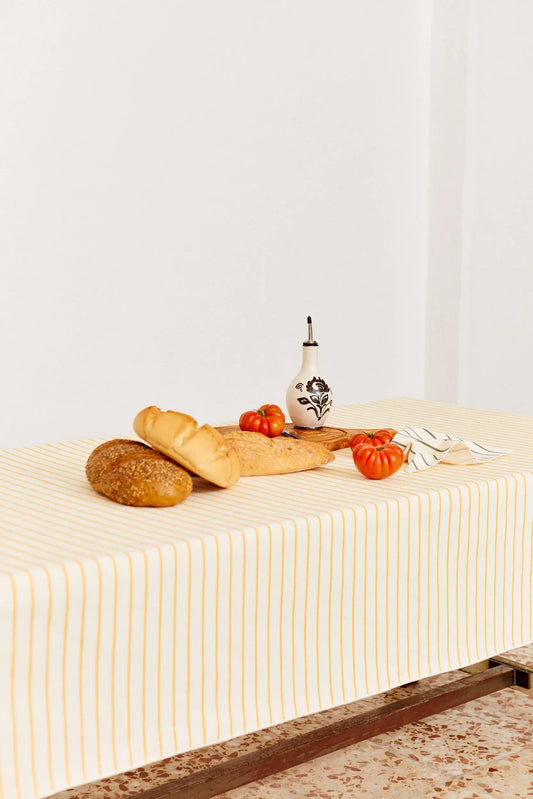 Mantel de tela color crema con finas rayas verticales amarillas, decorado con pan, tomates y una aceitera artesanal.