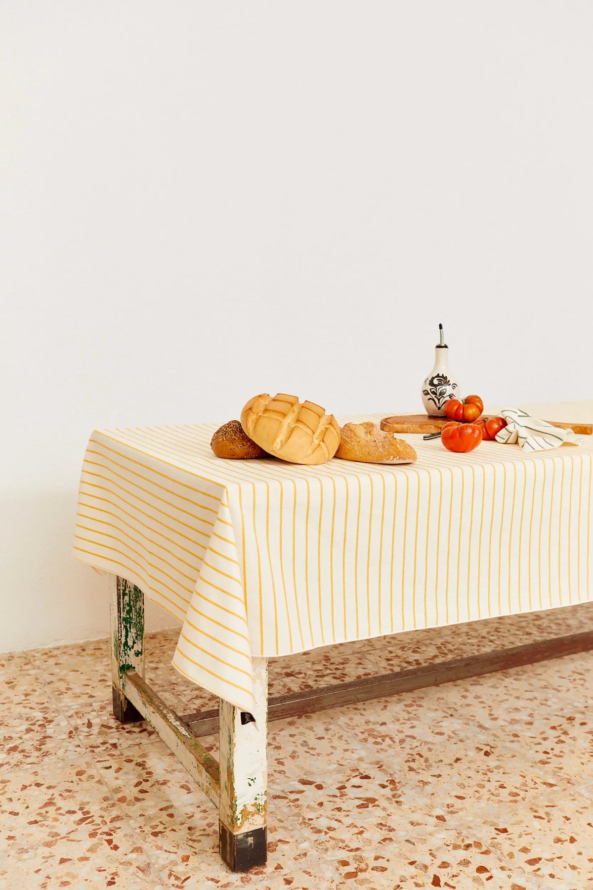 Mantel de tela blanco con rayas verticales amarillas, extendido sobre una mesa rústica con pan y tomates encima.