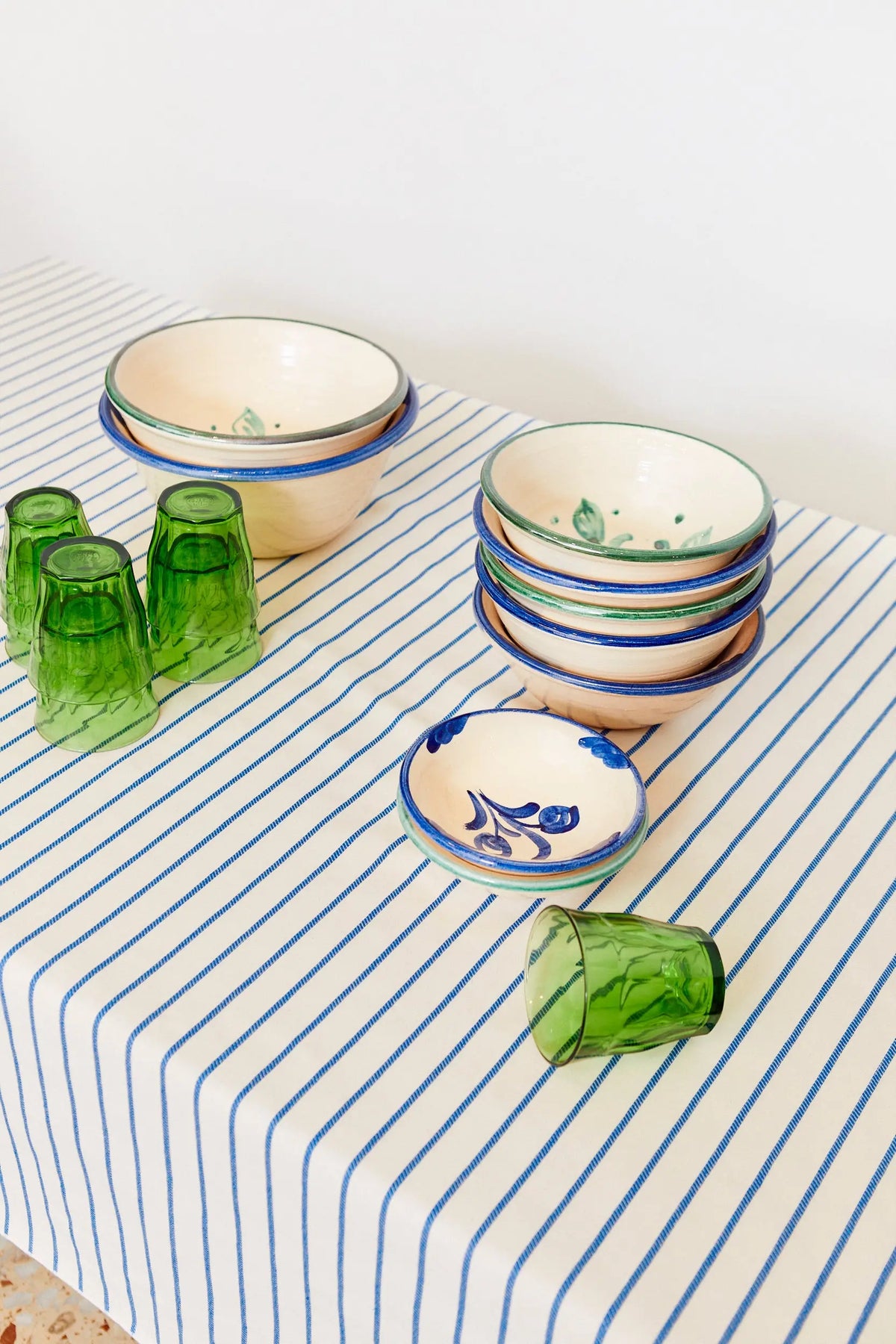 Mantel de tela blanco con finas rayas azules, sobre el que se disponen cuencos de cerámica y vasos de vidrio verde.