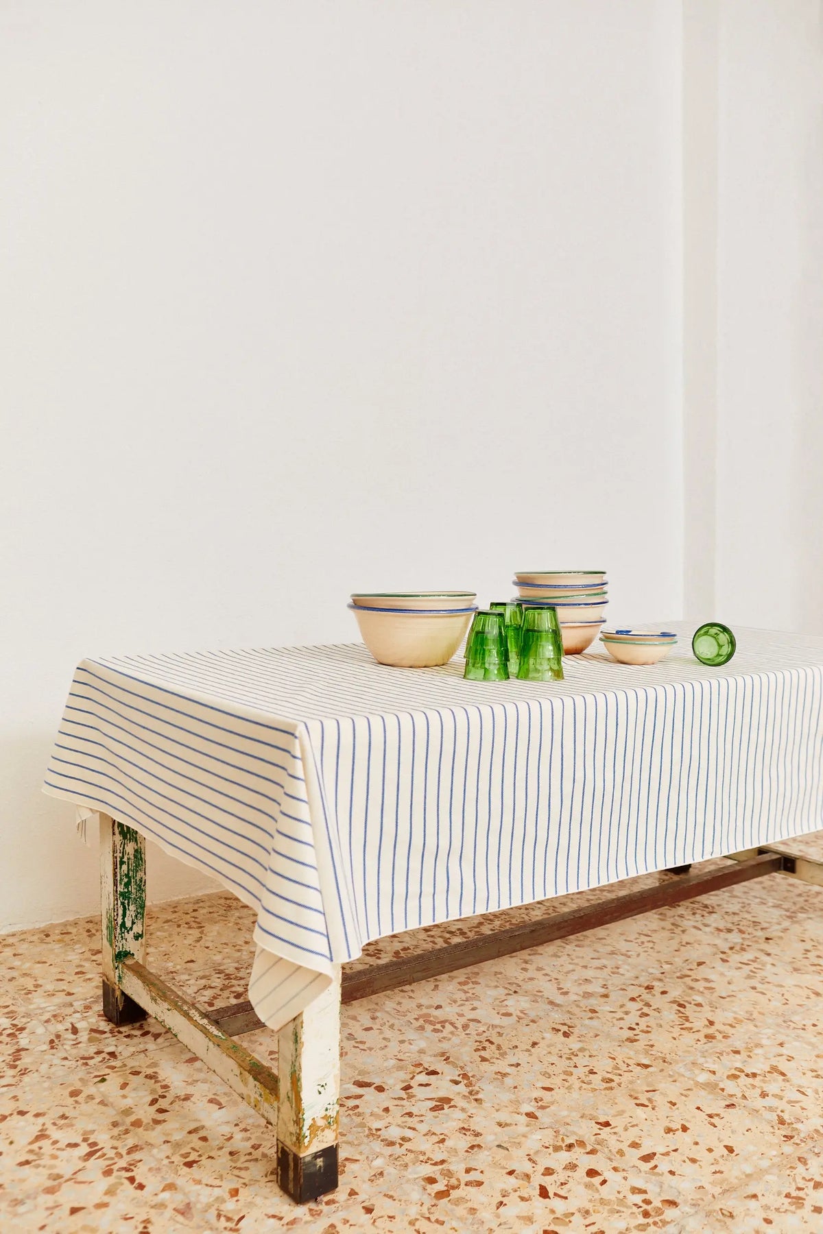 Mantel de algodón blanco con rayas azules verticales sobre una mesa de madera rústica con cuencos y vasos verdes.