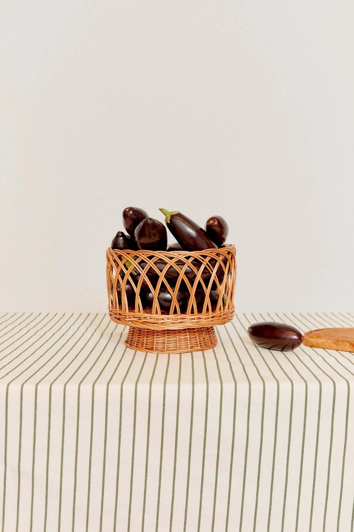 Tablecloth with a cream base and thin green vertical stripes, featuring a woven wicker basket filled with eggplants.