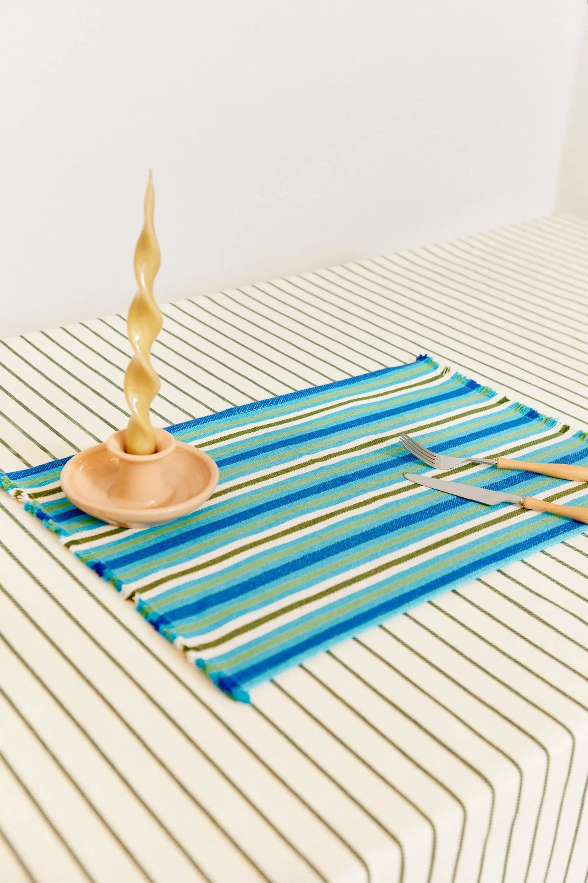 Mantel individual de tejido artesanal con rayas horizontales en tonos azules y verdes, con flecos y cubiertos sobre mesa.