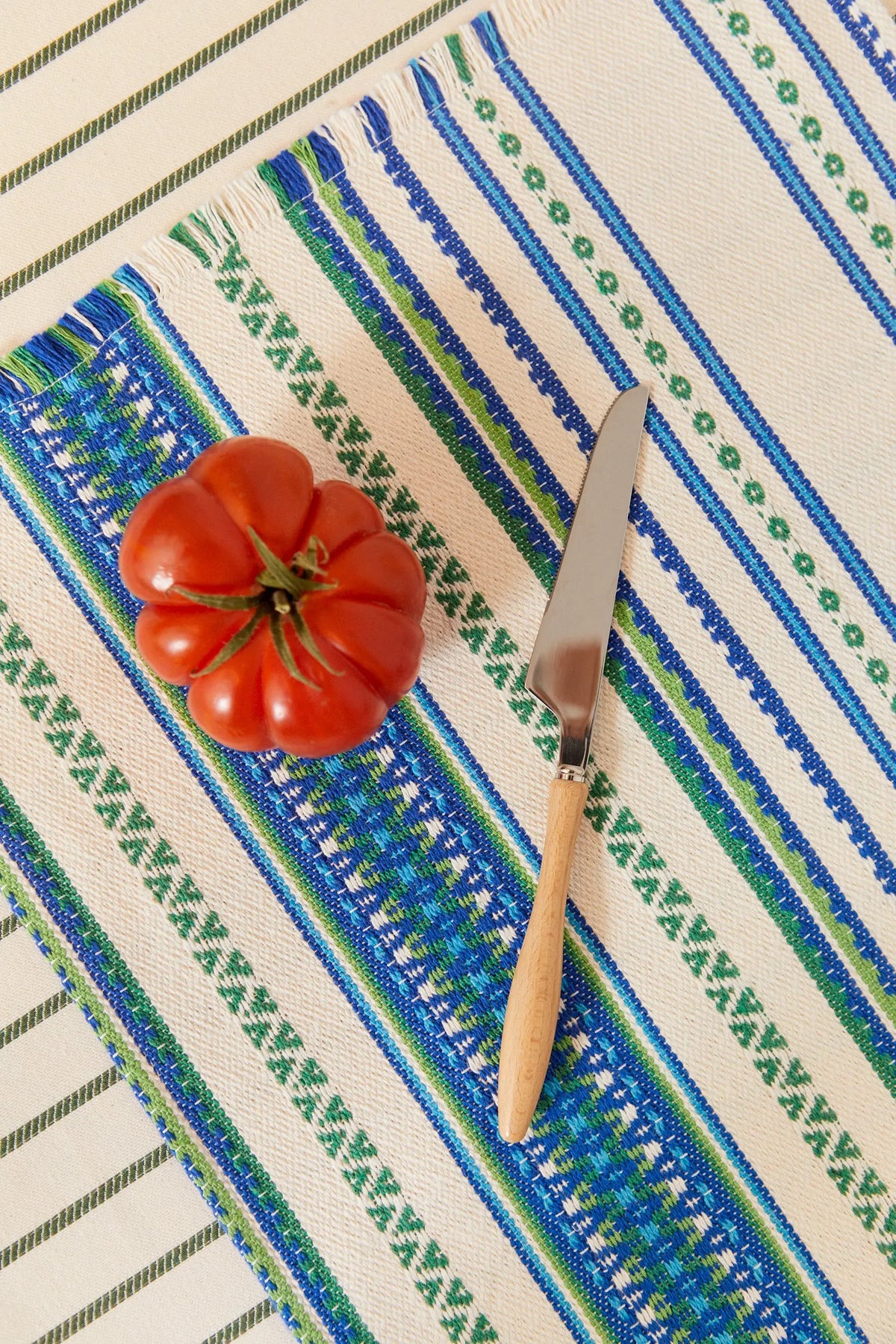 Pack de dos manteles individuales de algodón tejido con rayas y cenefas en tonos azules y verdes, con tomate y cuchillo.