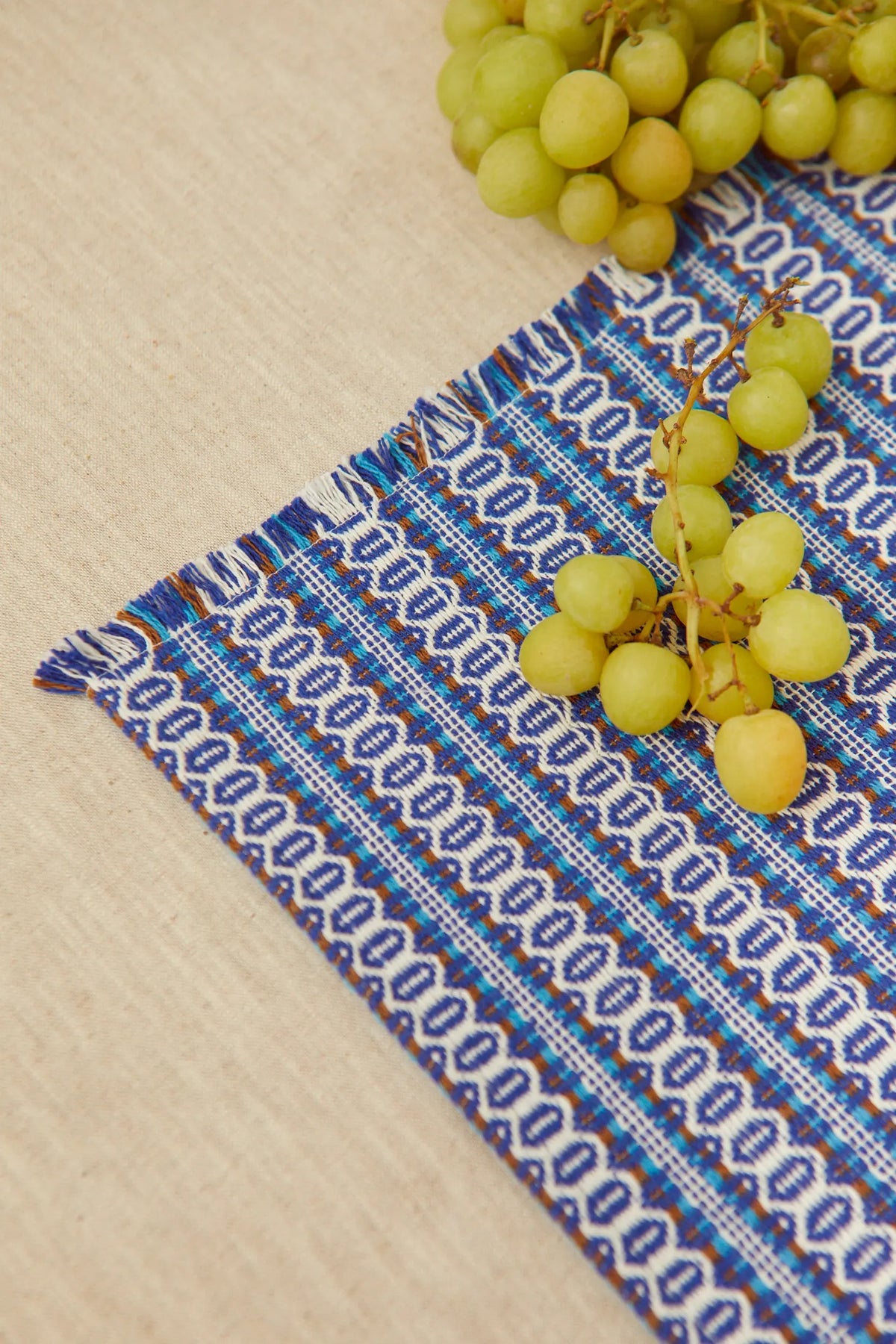 Mantel individual retro azul con patrón ojo de perdiz, textura tejida con flecos y racimo de uvas frescas encima.