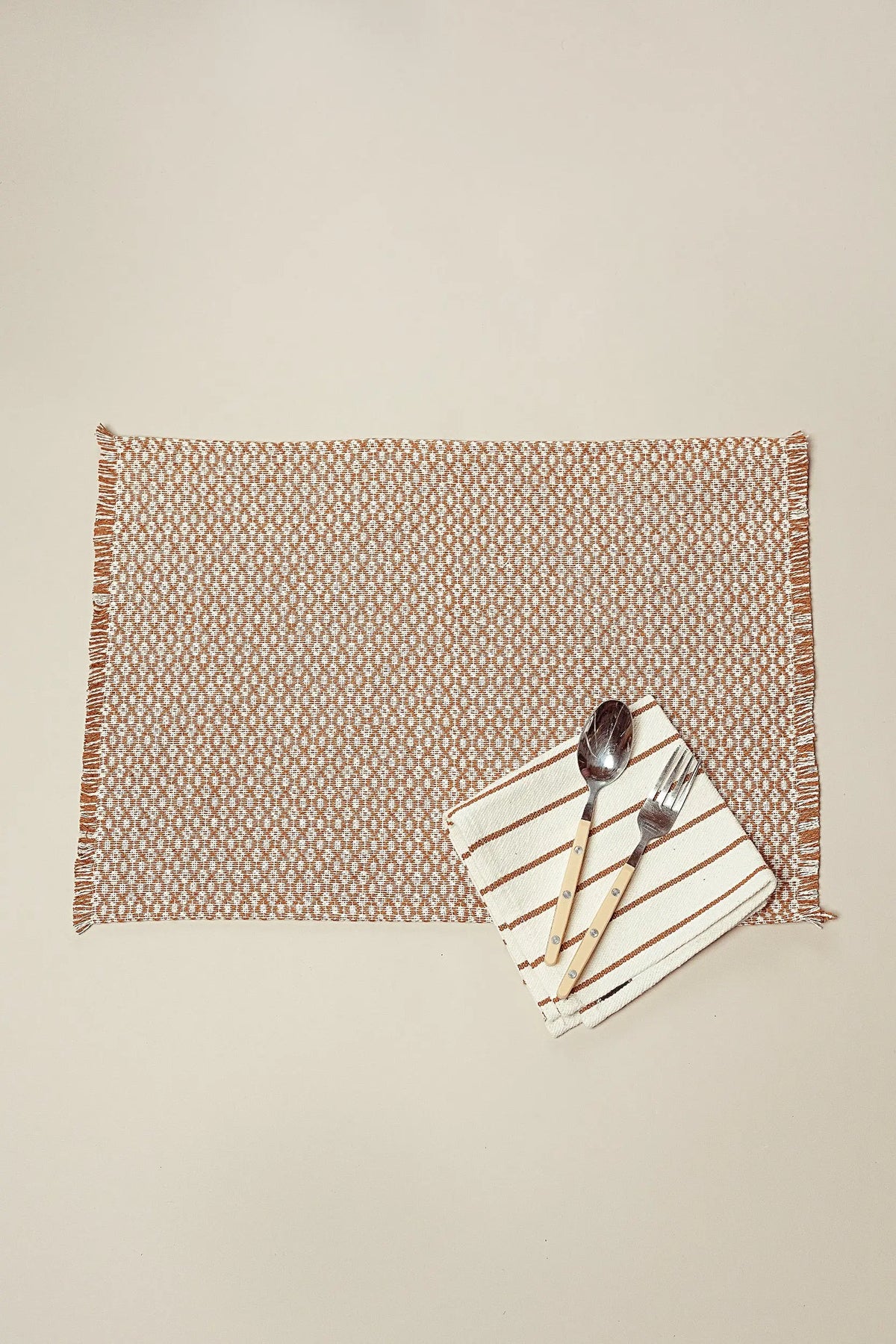 Caramel and white patterned placemat with fringed edges, featuring a striped napkin, spoon, and fork on top.