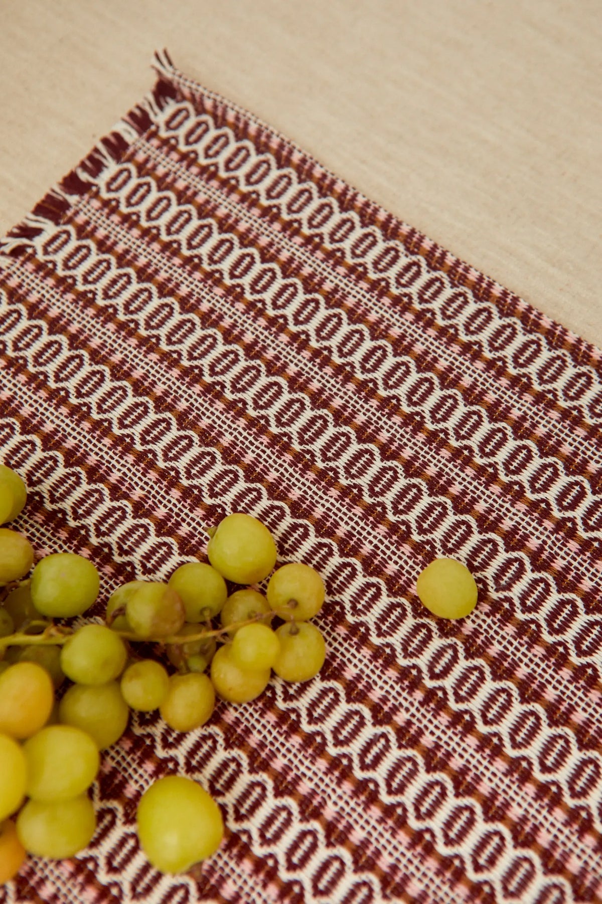 Mantel individual estilo retro con patrón ojo de perdiz granate y crema sobre fondo beige, con racimo de uvas encima.