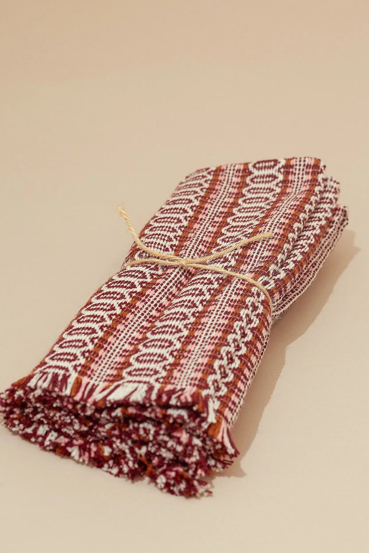 Two folded placemats with a maroon, white, and copper woven pattern, fringed edges, tied with rustic string.