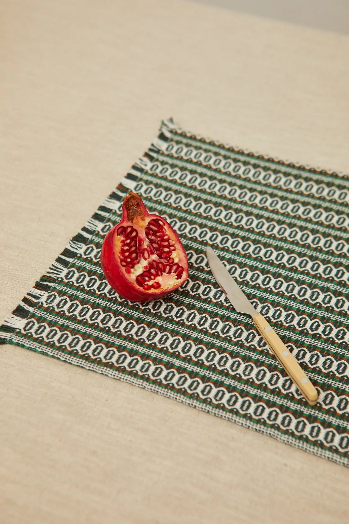 Rectangular green retro patterned placemat with fringed edges, featuring a cut pomegranate and a knife on top.