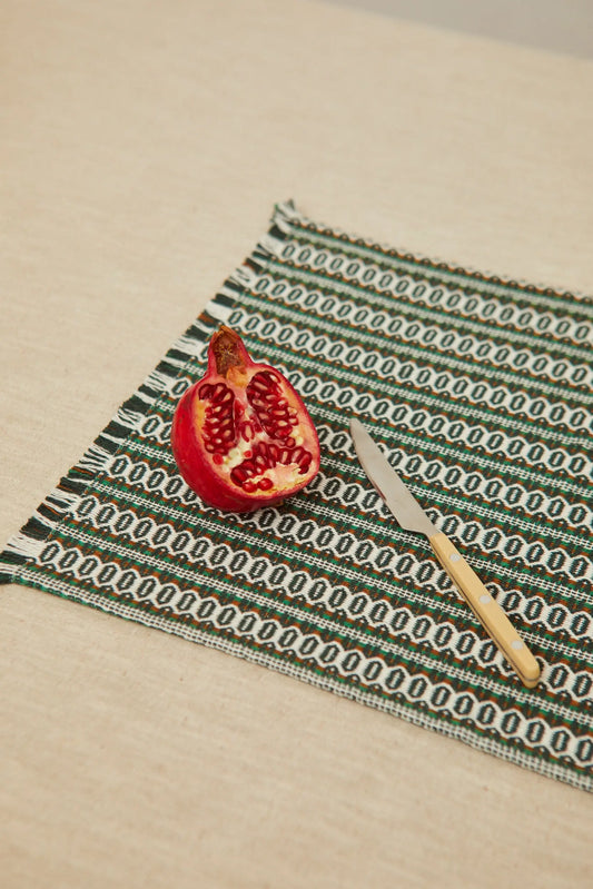 Rectangular green retro patterned placemat with fringed edges, featuring a cut pomegranate and a knife on top.