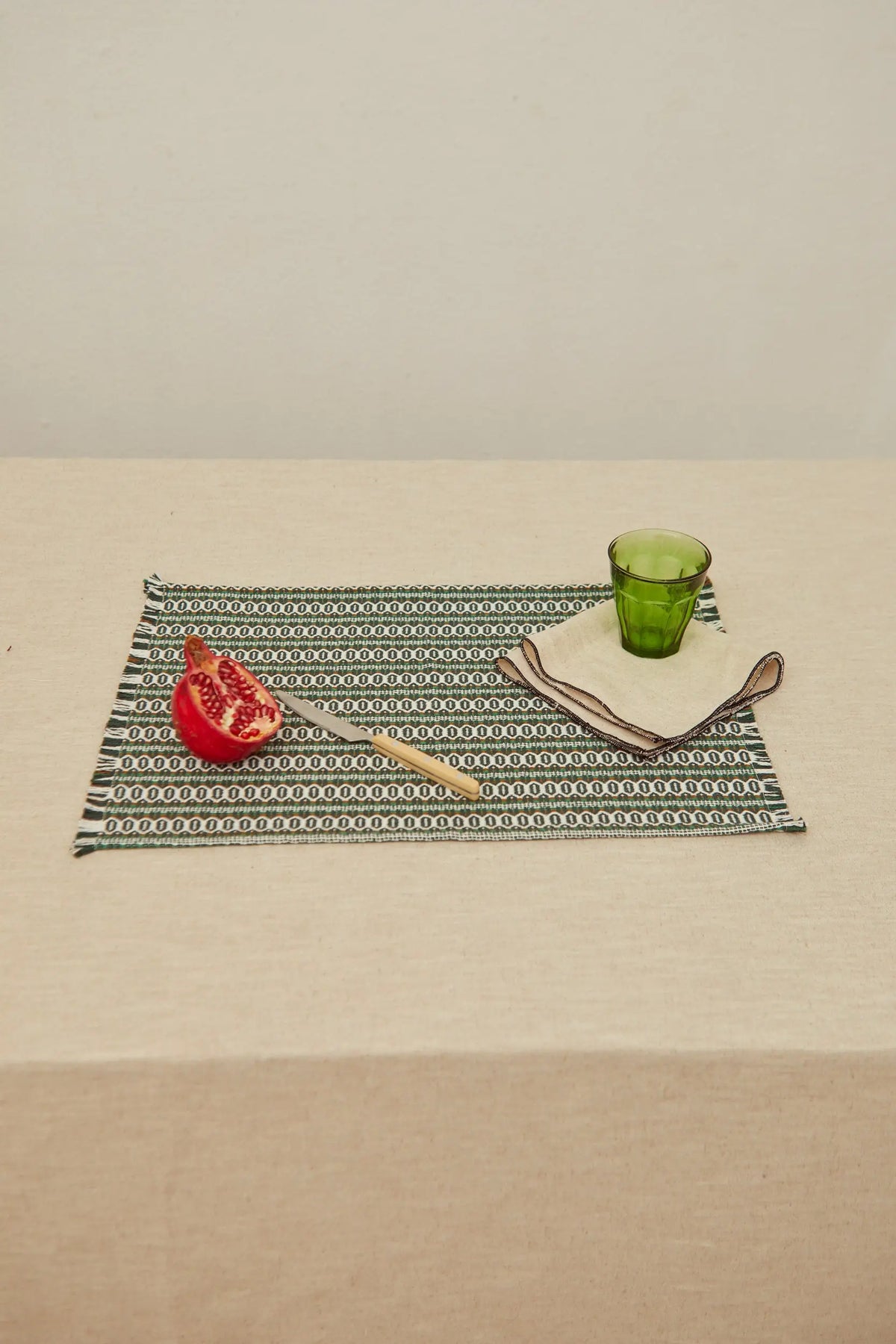 Rectangular green woven placemat with retro patterns, set with a pomegranate, knife, napkin, and green glass.