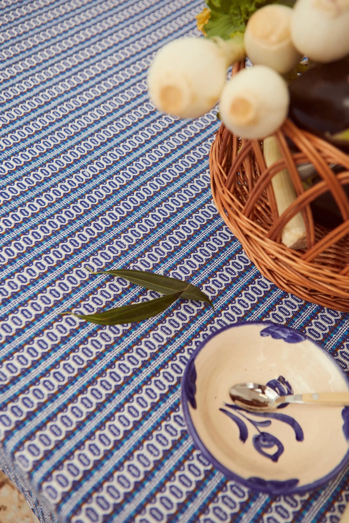 Mantel textil con patrón retro de ojo de perdiz en tonos azules, sobre mesa con cesta de mimbre y cuenco de cerámica.