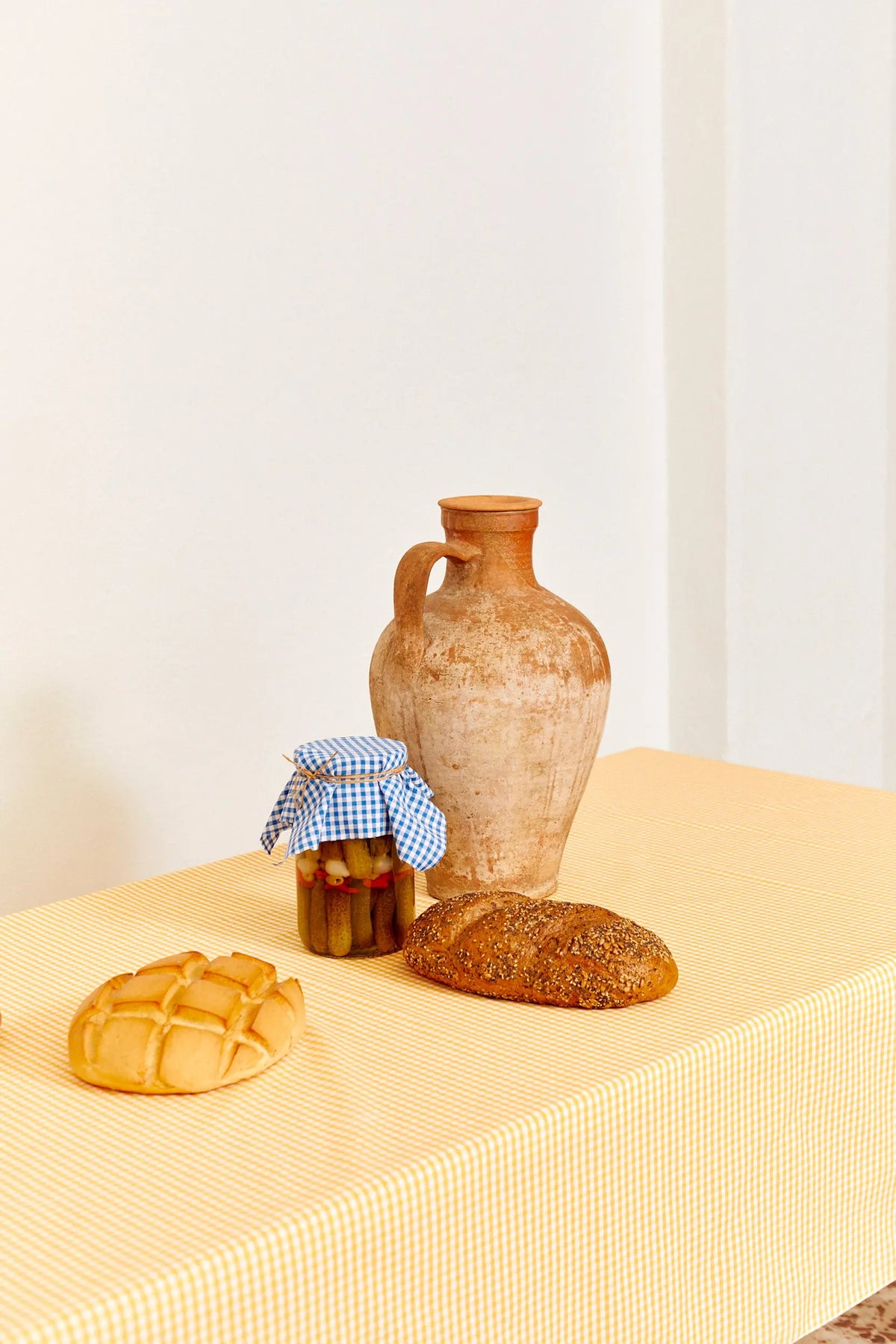 Mantel de tela con estampado de cuadros vichy amarillos sobre una mesa con pan y un jarrón de barro.