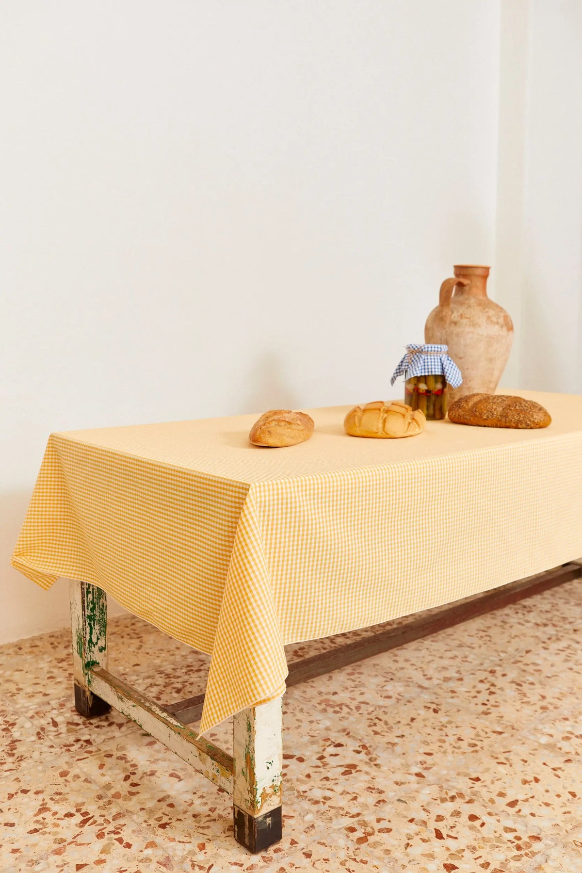 Mantel de tela con estampado de cuadros vichy en color amarillo sobre una mesa rústica con pan y un jarrón de barro.