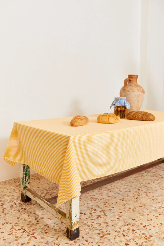 Mantel de tela con estampado de cuadros vichy en color amarillo sobre una mesa rústica con pan y un jarrón de barro.