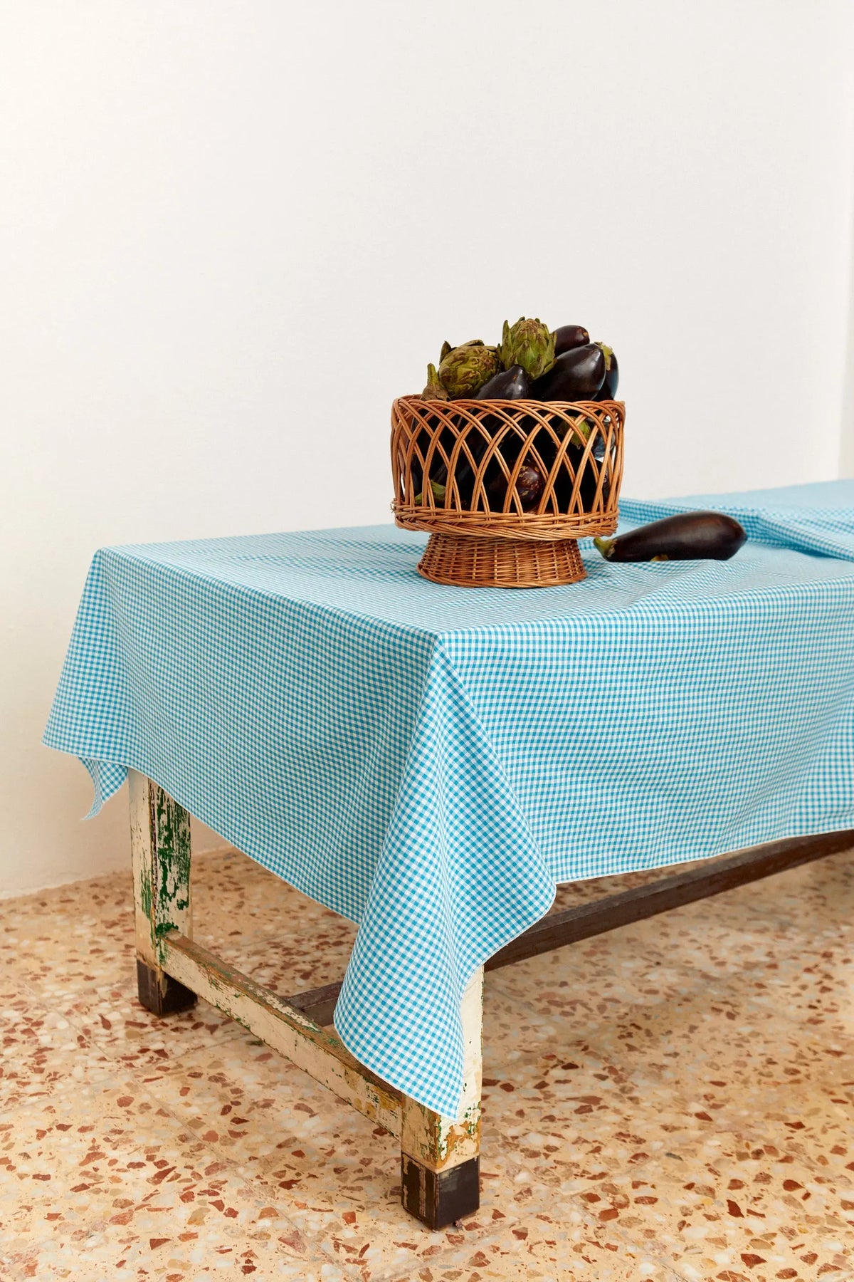 Mantel de tela con estampado de cuadros vichy en color celeste sobre una mesa rústica con una cesta de berenjenas.