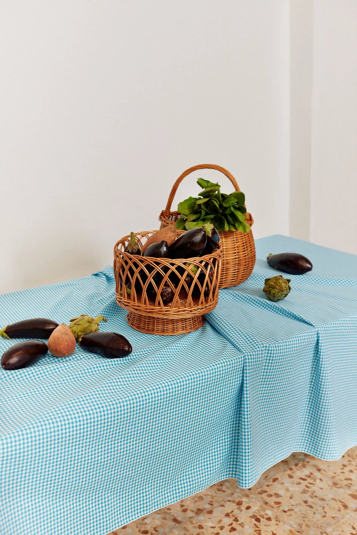 Mantel de tela con estampado de cuadros vichy en color celeste, sobre una mesa con cestas de mimbre y verduras frescas.