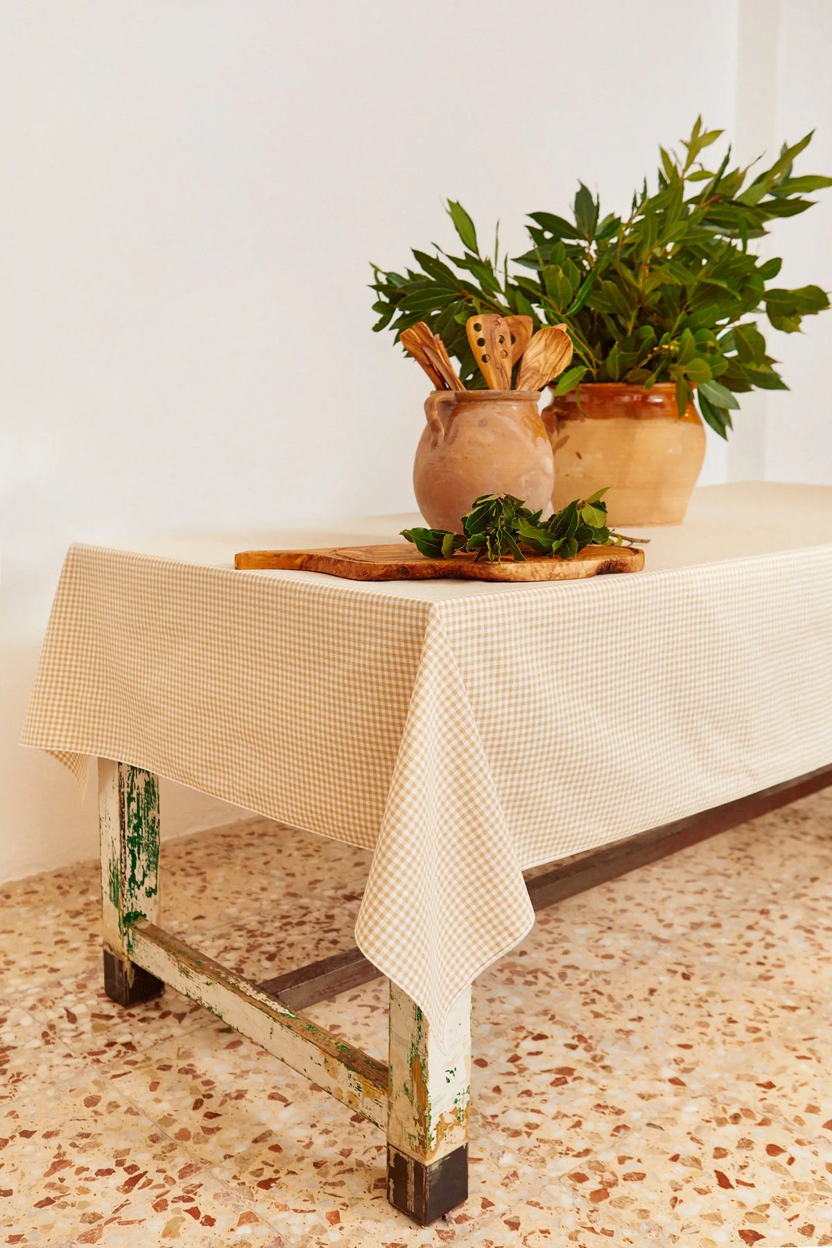 Mantel de algodón con estampado de cuadros vichy en color beige sobre una mesa rústica con utensilios y plantas.