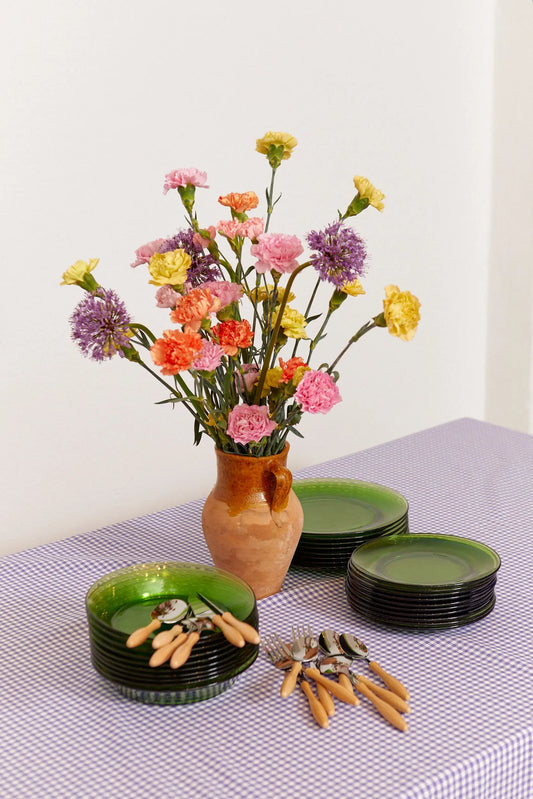 Mantel de cuadros vichy color malva con jarrón de flores, platos verdes y cubiertos con mango de madera sobre la mesa.