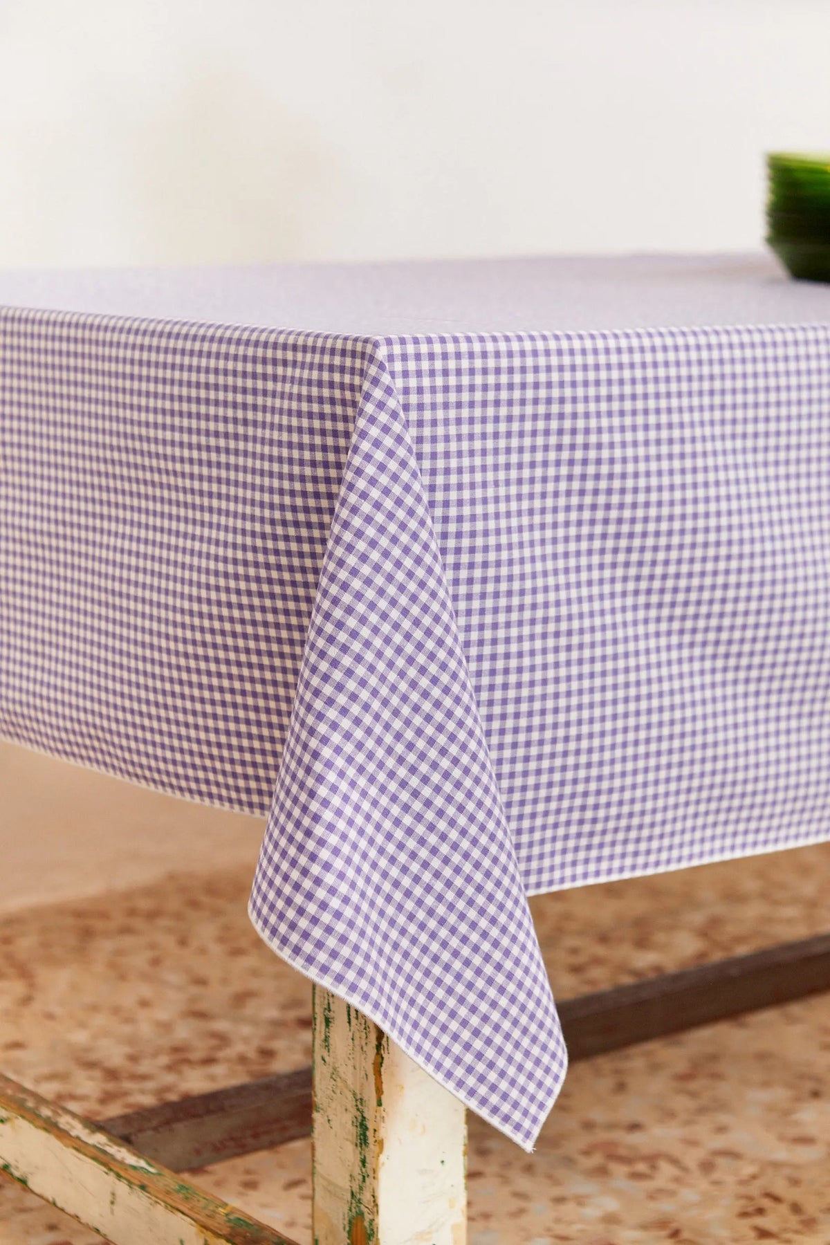 Mantel textil rectangular con estampado de cuadros vichy en color malva y blanco, cubriendo una mesa de estilo rústico.
