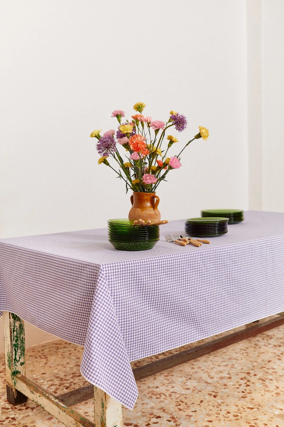Mantel de algodón con estampado de cuadros vichy malva sobre una mesa con flores y vajilla de cristal verde.