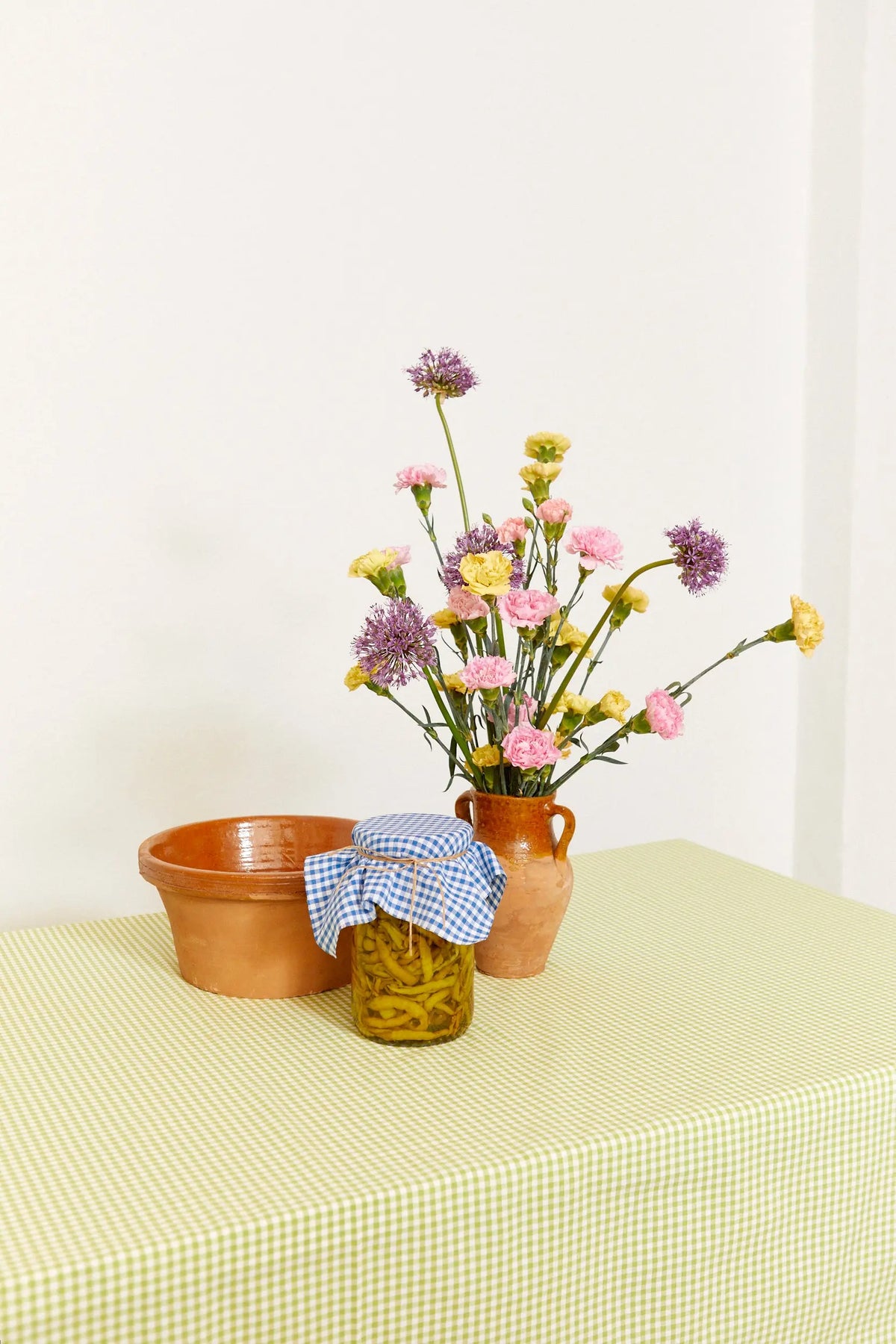 Mantel de cuadros vichy verde sobre mesa con jarrón de flores y frasco decorado. Estilo rústico y acogedor.