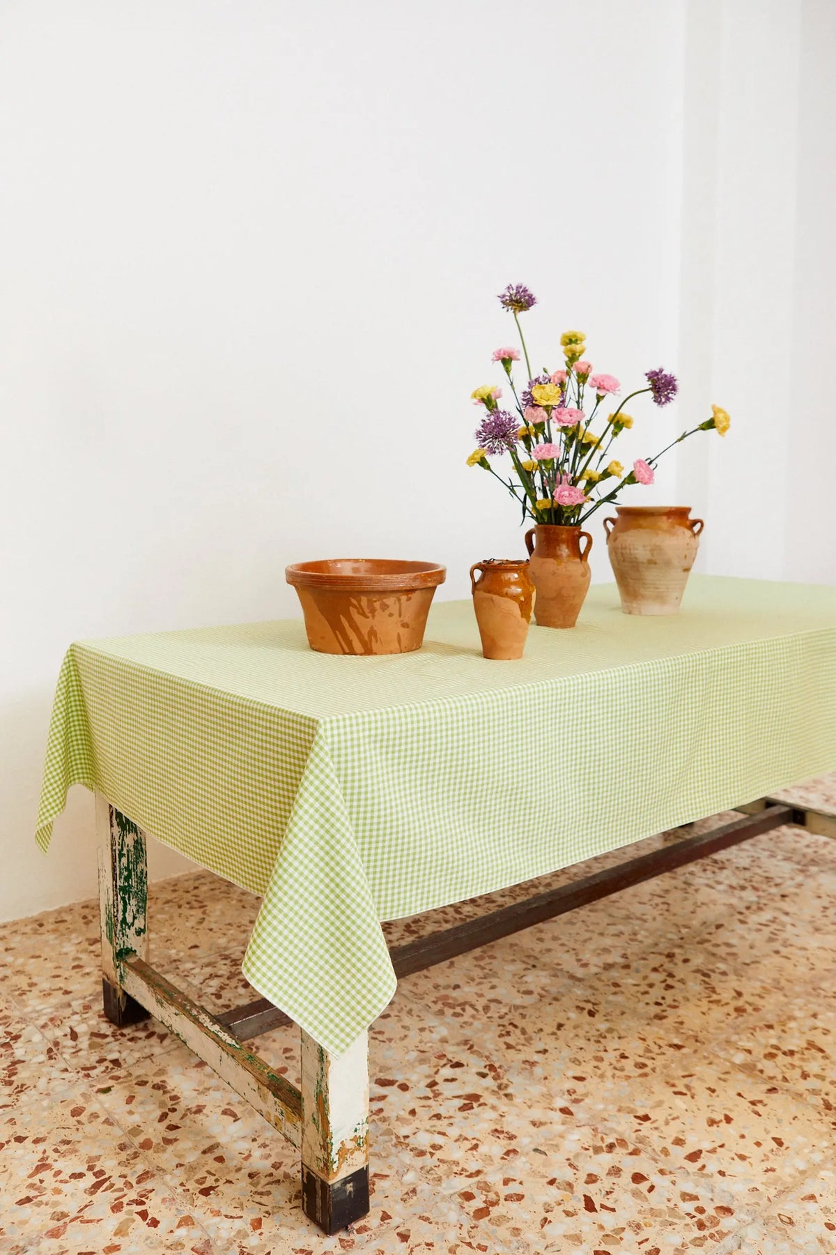 Mantel de tela con estampado de cuadros vichy en color verde claro sobre una mesa rústica con vasijas y flores.