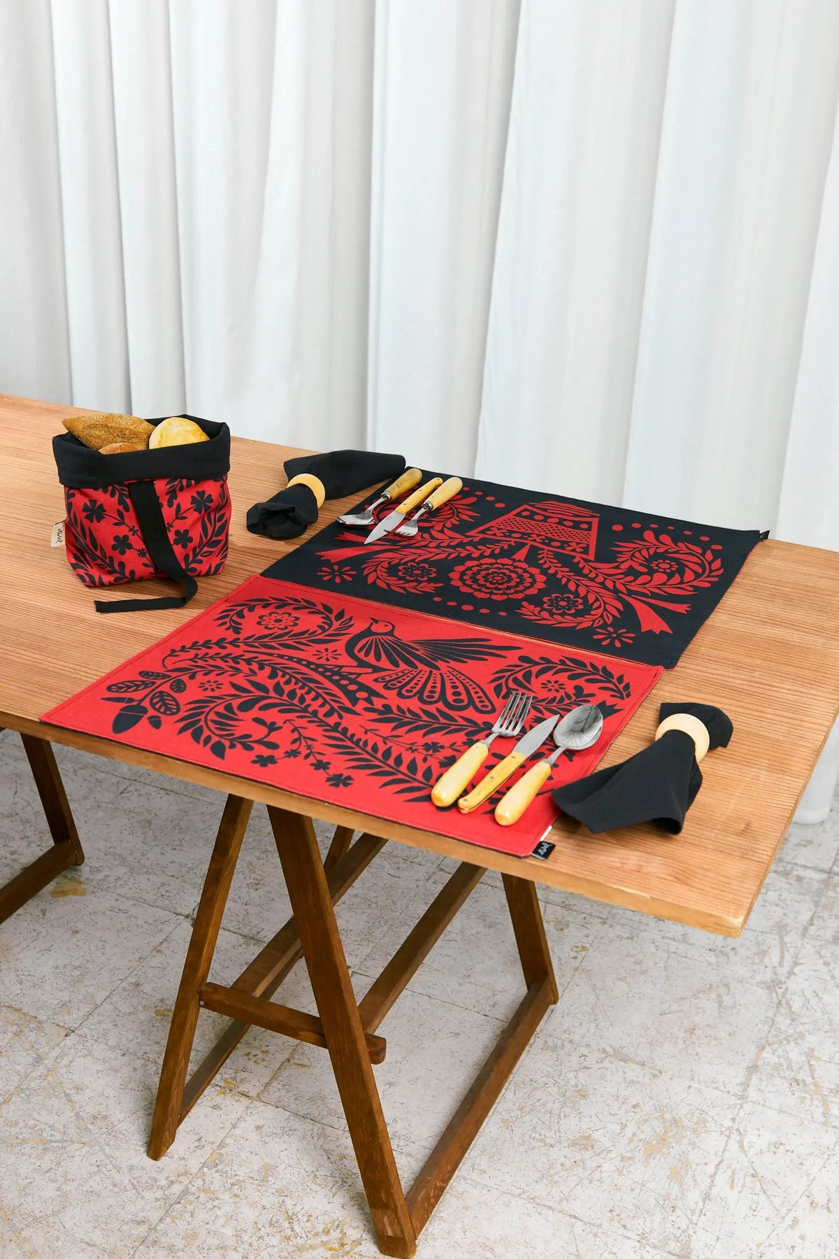 Small fabric bread basket with black floral print on red, placed on a wooden table with matching table settings.
