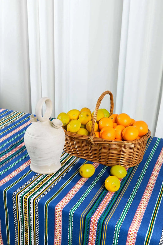 Mantel de tela con rayas multicolores sobre fondo azul, con una cesta de mimbre llena de limones y naranjas y un botijo.