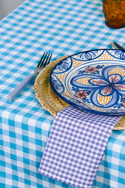 Purple gingham cloth napkin on a table set with a blue checkered tablecloth, wicker charger, and ornate plate.