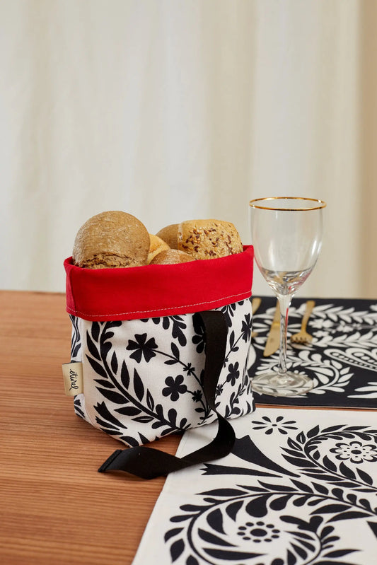 Panera de tela con estampado floral blanco y negro, borde superior rojo, asa negra y pan en su interior.