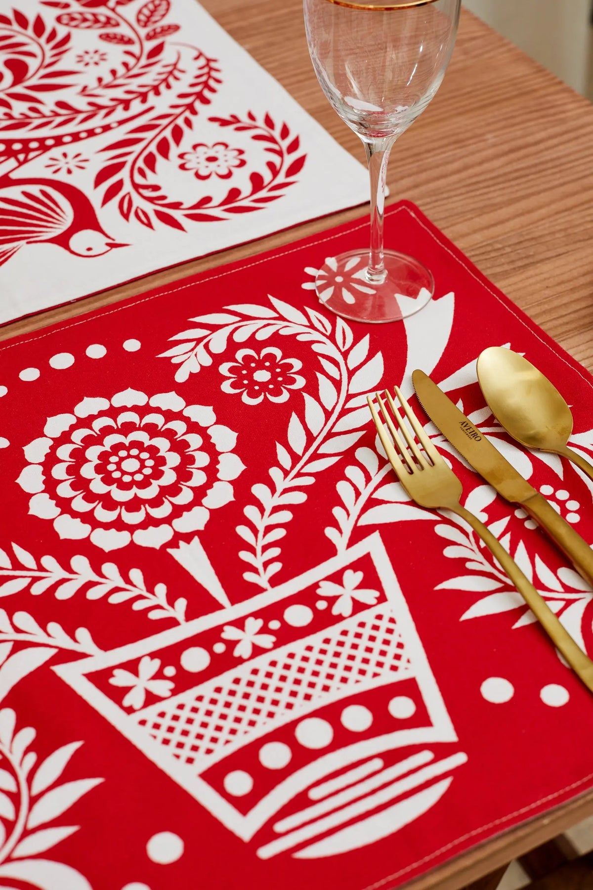 Two reversible placemats featuring a red and white folk-art floral pattern on a wooden table with gold cutlery.