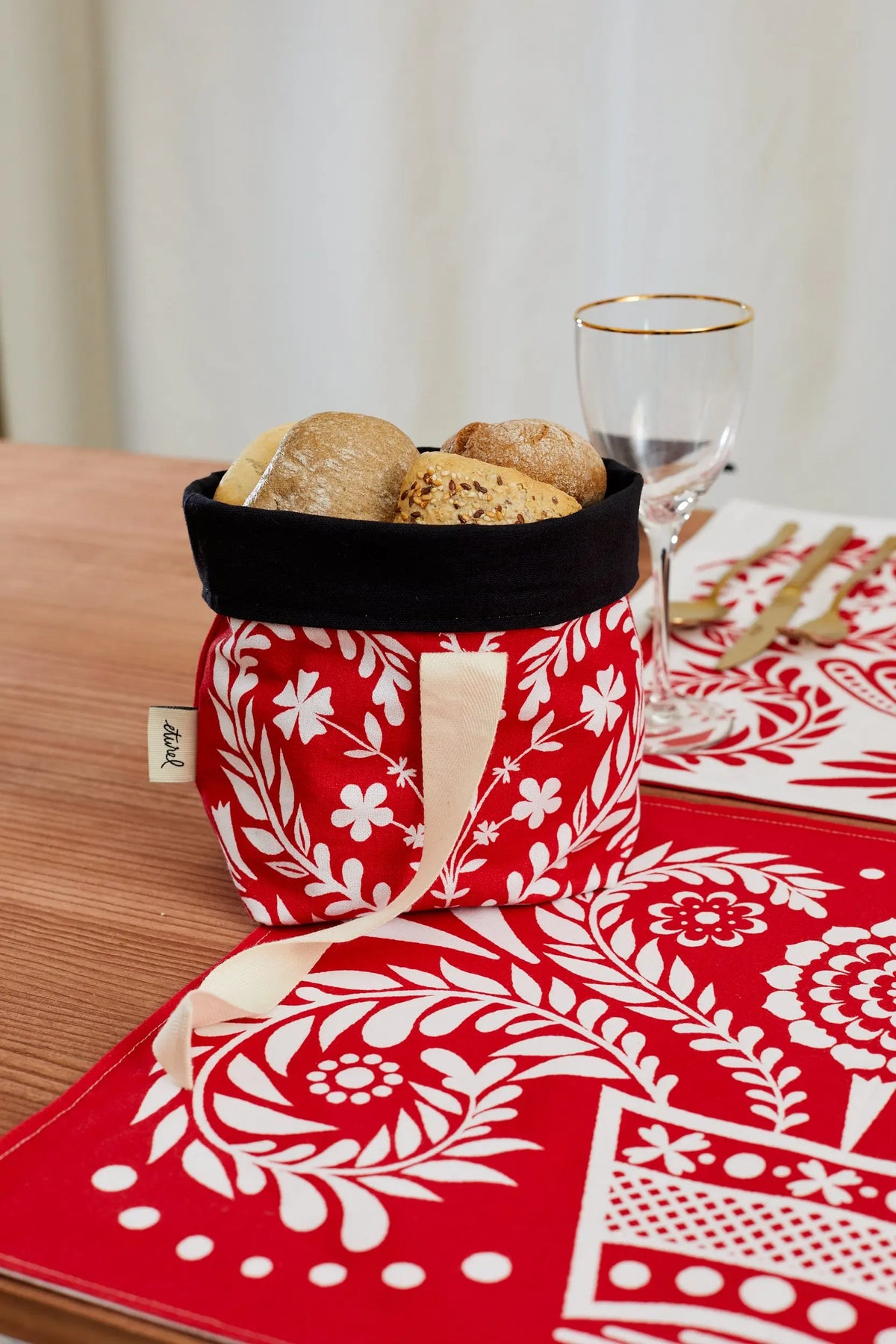 Panera de tela roja con estampado floral blanco y borde interior negro, con panecillos dentro sobre mesa de madera.