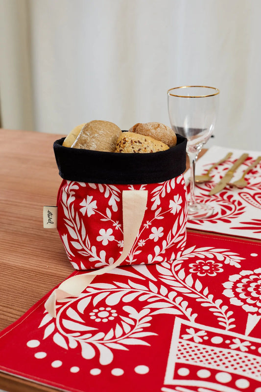 Panera de tela roja con estampado floral blanco y borde interior negro, con panecillos dentro sobre mesa de madera.