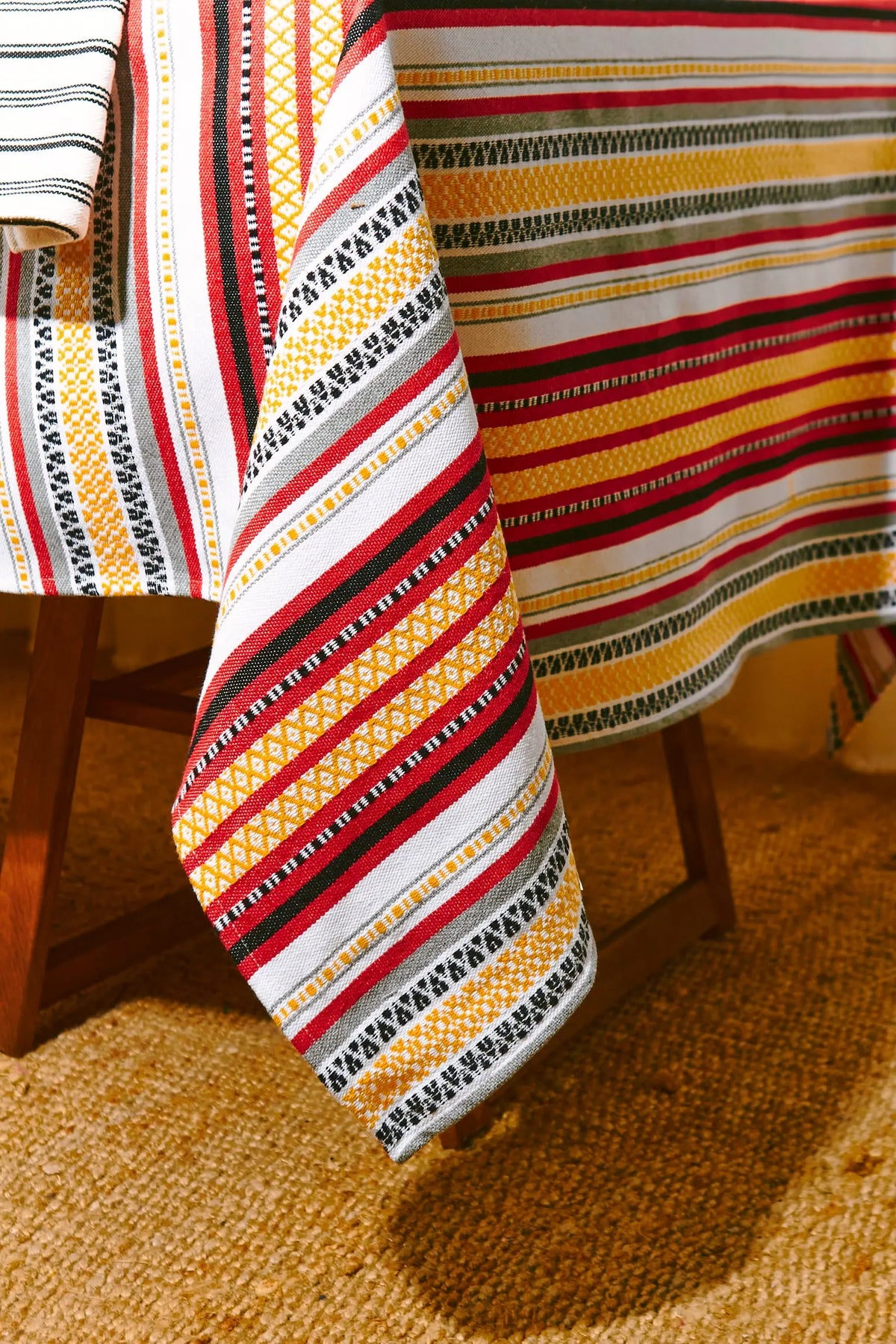 Mantel de tela con estampado de franjas multicolores en rojo, amarillo, gris y negro sobre una mesa de madera.
