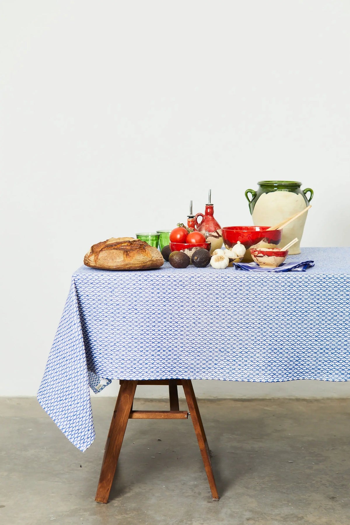 Blue and white bird's eye pattern tablecloth on a wooden table set with artisan ceramics, bread, and vegetables.