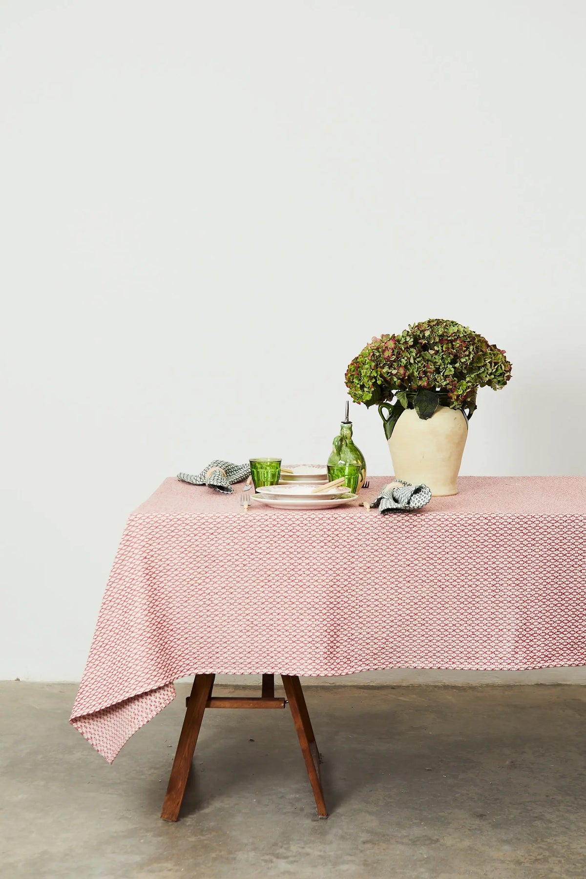 Maroon and white bird's-eye pattern tablecloth on a wooden table with simple place settings and a floral vase.