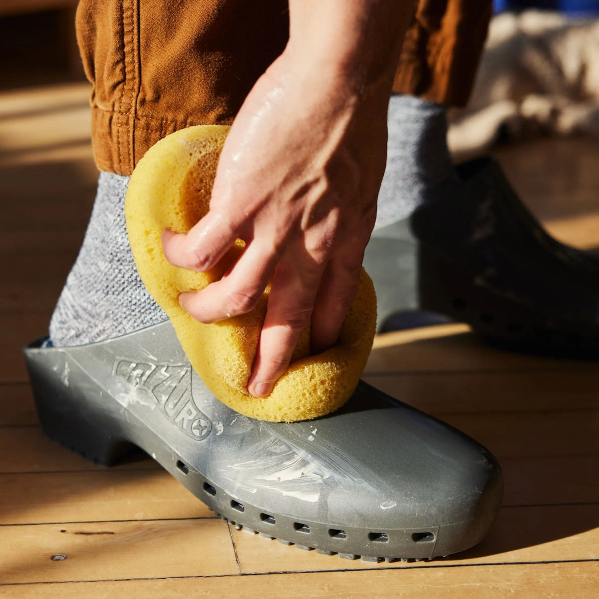 Metallic grey slip-on clog being cleaned with a yellow sponge, showing a smooth, solid design without top holes.