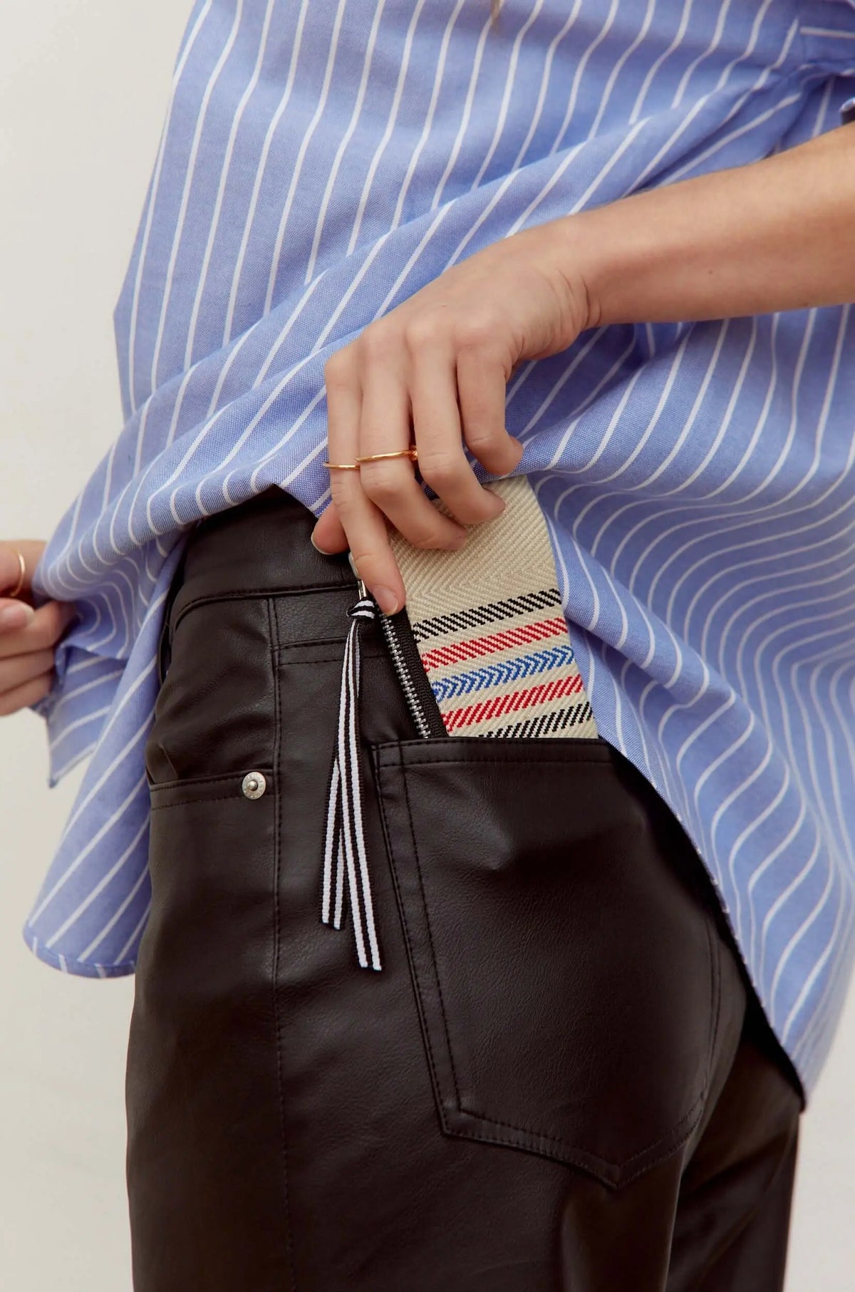 Monedero de sarga color crudo con rayas rojas y azules, cremallera negra y tirador de tela, guardado en bolsillo trasero.