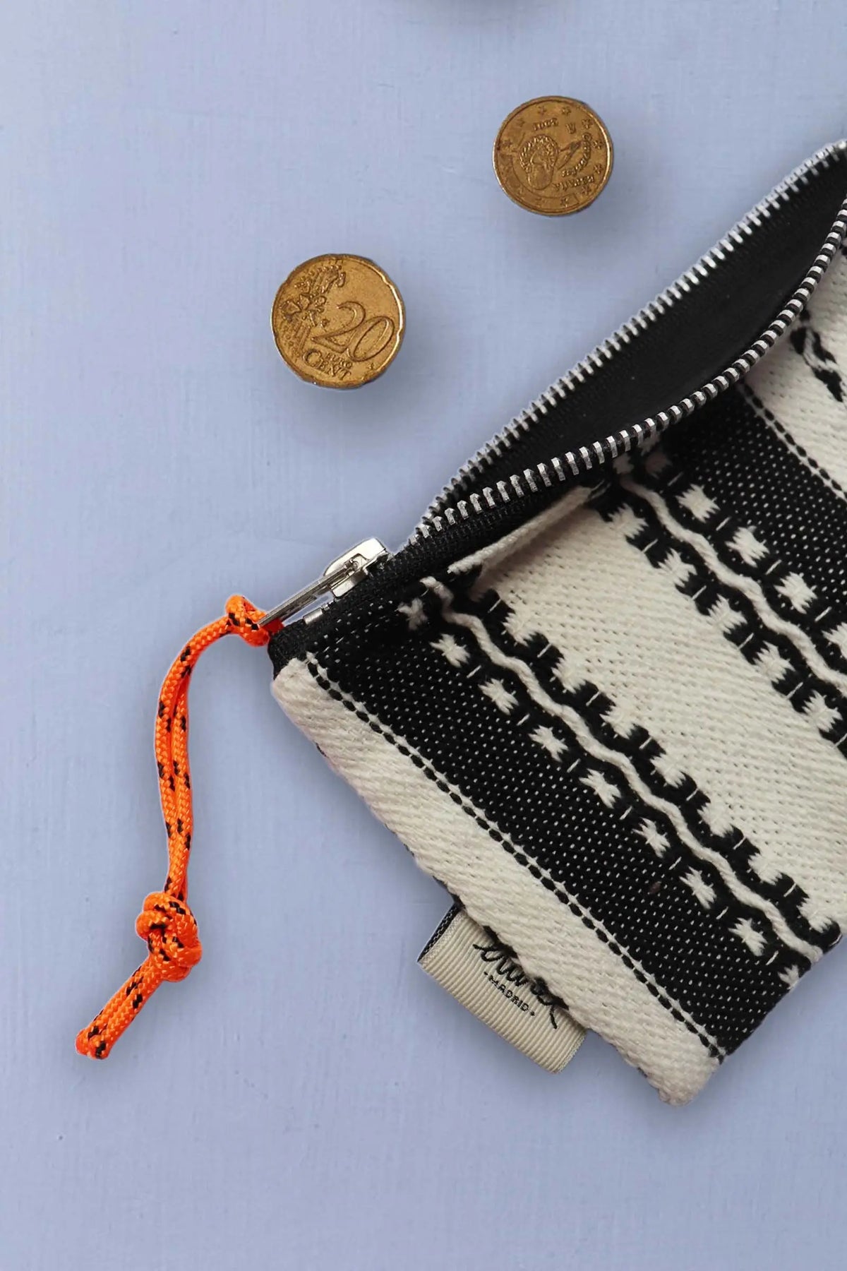 Monedero de tela tejida estilo alpujarra en blanco y negro con cremallera y tirador naranja junto a dos monedas.