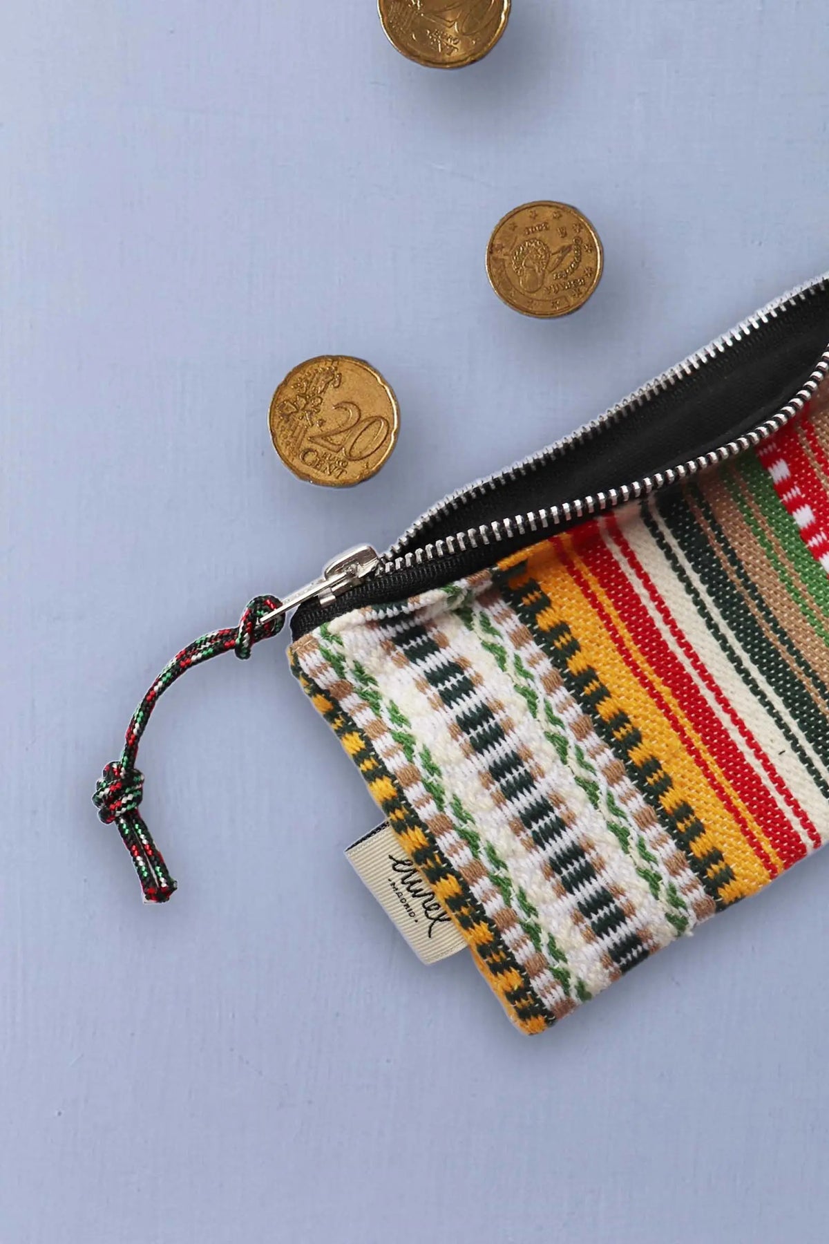 Monedero artesanal con tejido de rayas multicolores, cremallera metálica y tres monedas de euro sobre fondo gris claro.