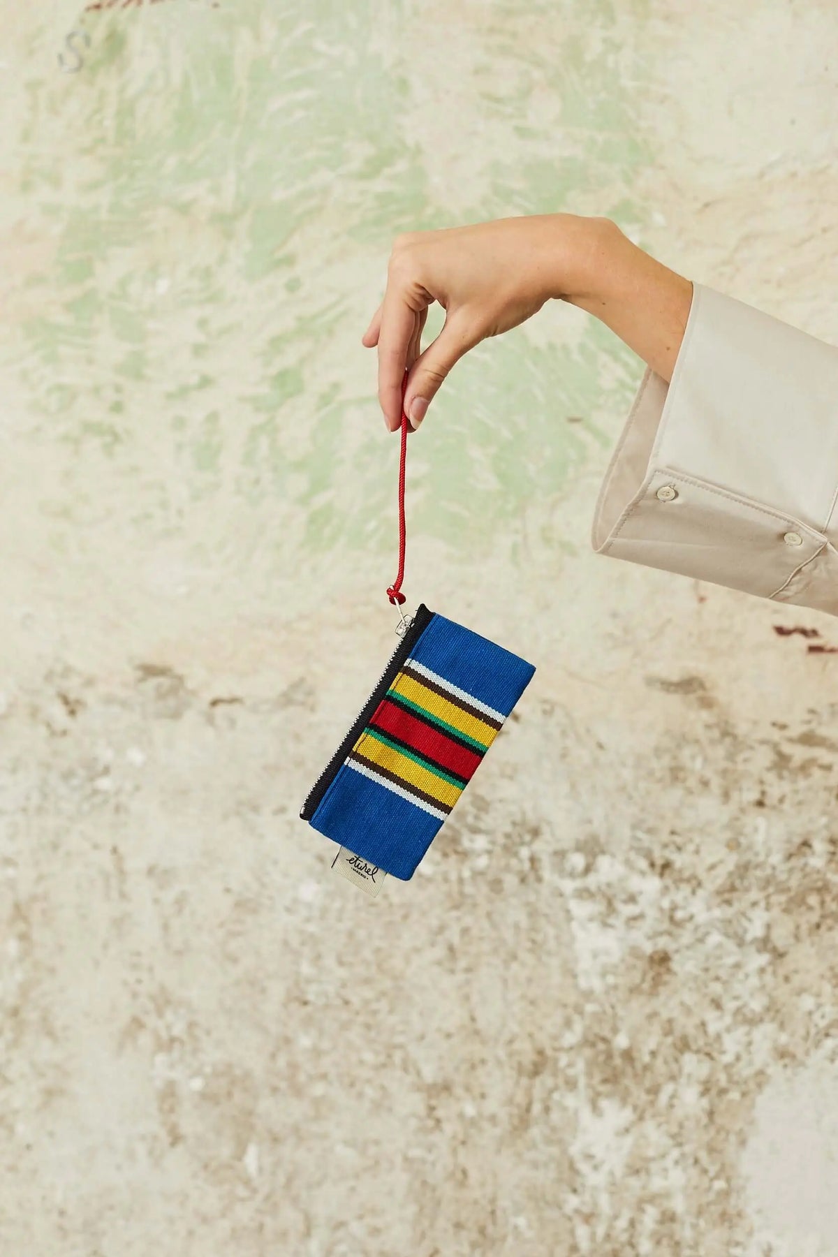 Monedero de lona estilo hamaca con franjas azules, rojas y amarillas, colgando de una cuerda roja por una mano.