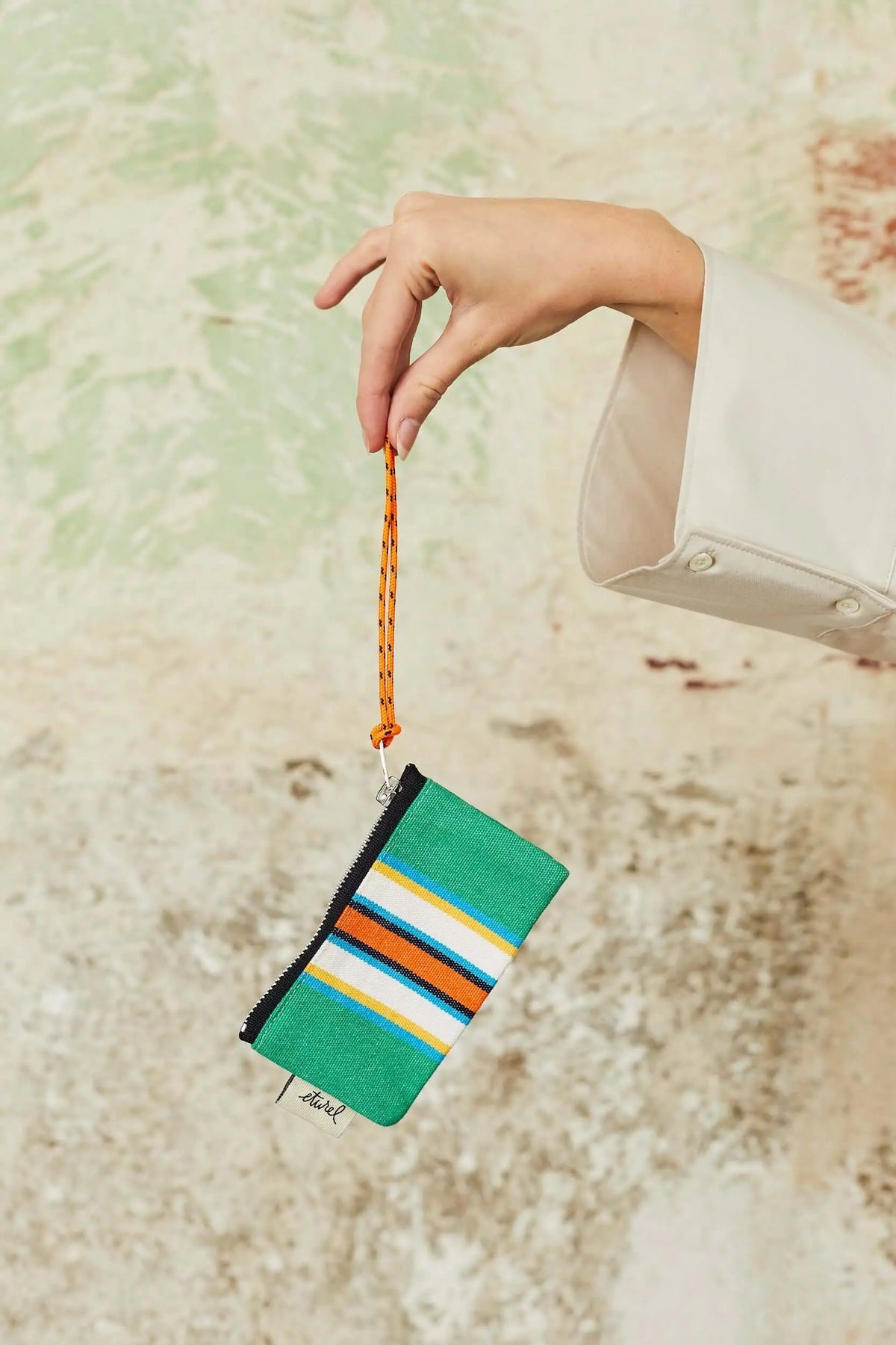 Monedero de lona estilo hamaca en verde a rayas con cremallera negra y cordón naranja, sostenido por una mano.