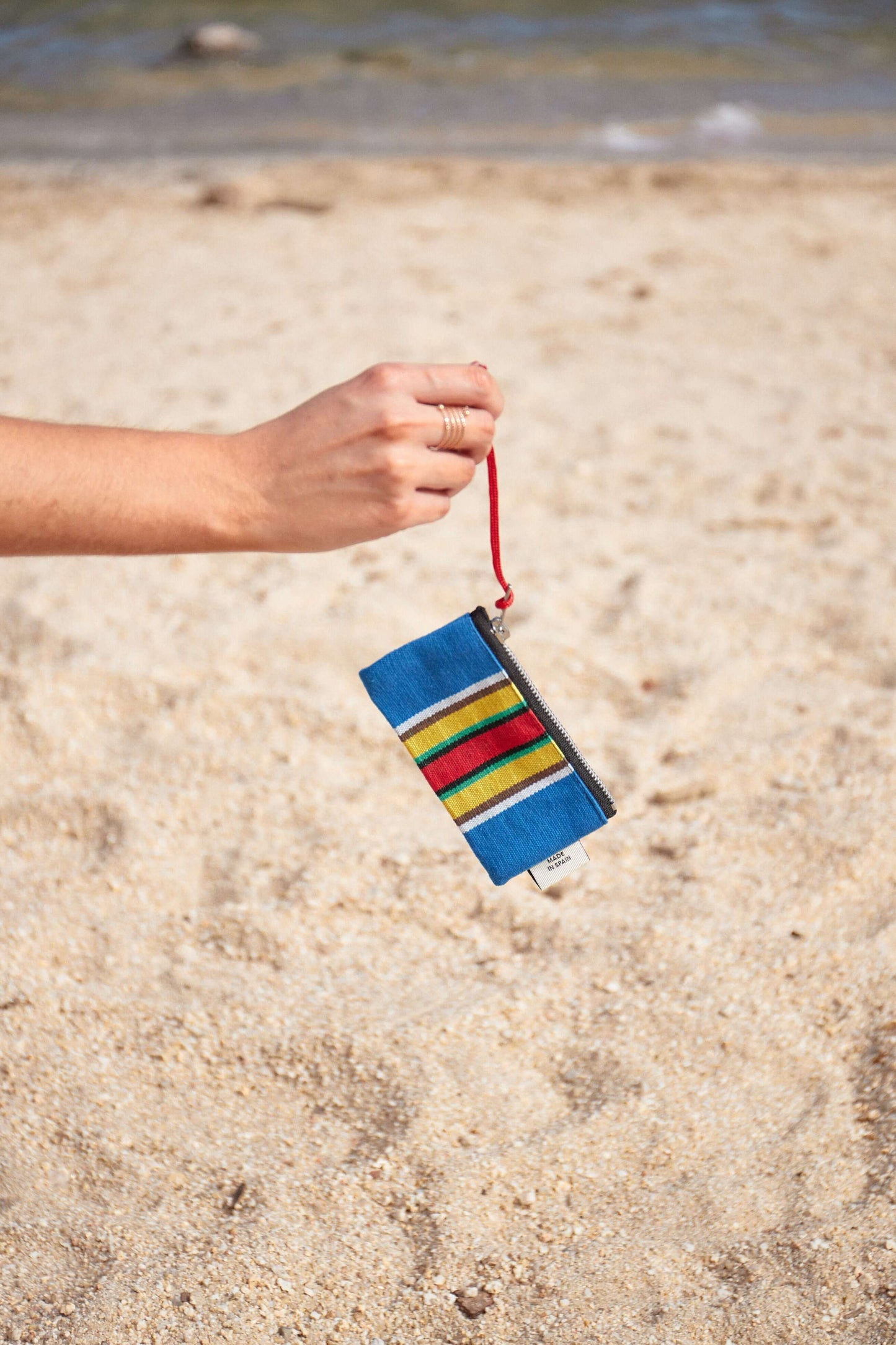 Blue canvas zippered coin purse with colorful horizontal stripes, held by a red cord against a beach background.