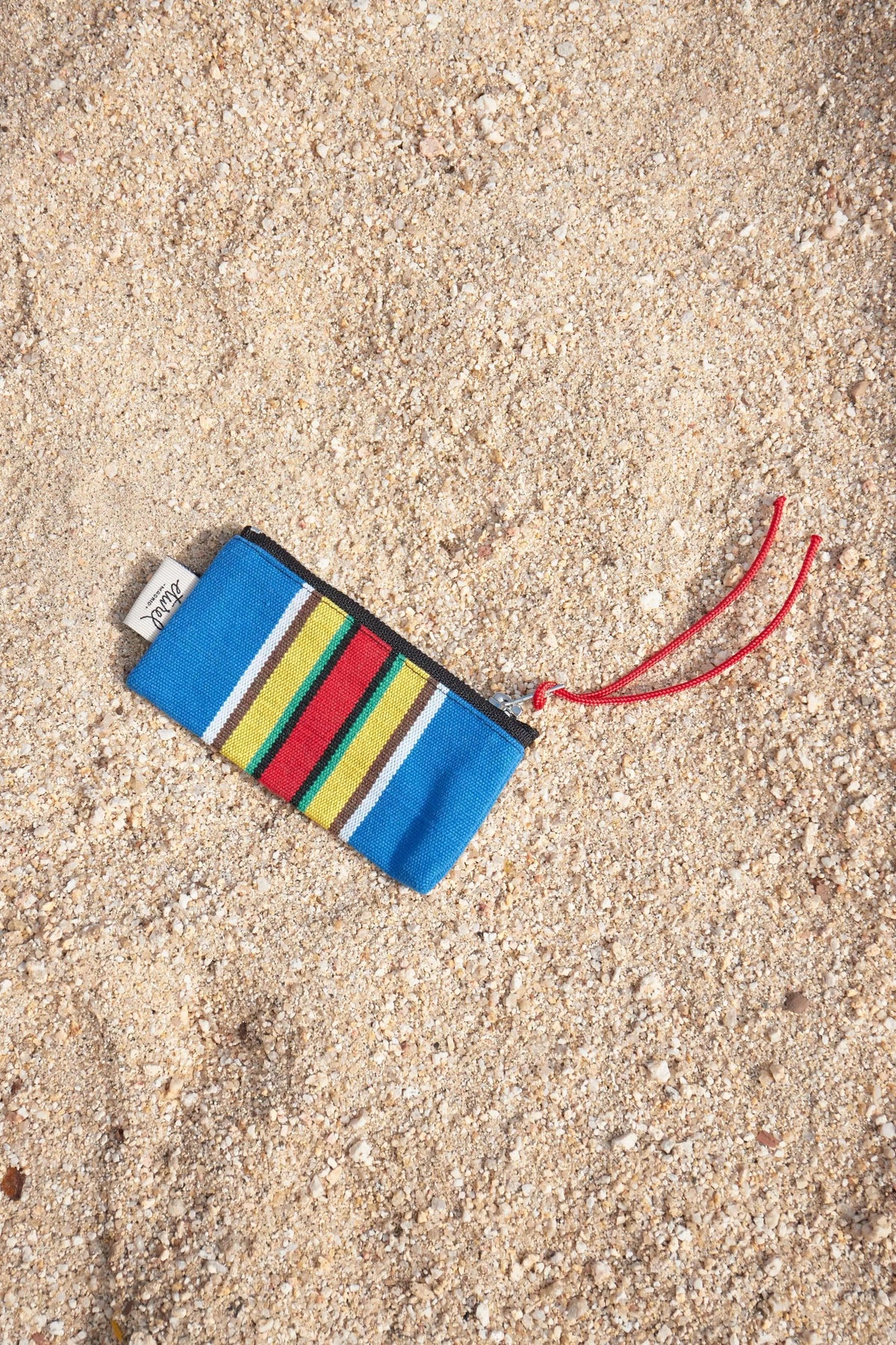 Monedero de lona estilo hamaca con rayas azules, amarillas y rojas, cremallera negra y tirador rojo sobre arena.