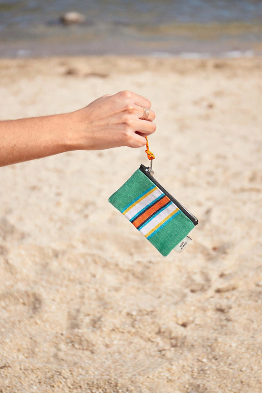 Monedero de lona verde a rayas de colores con cierre de cremallera, sostenido por una mano en una playa.