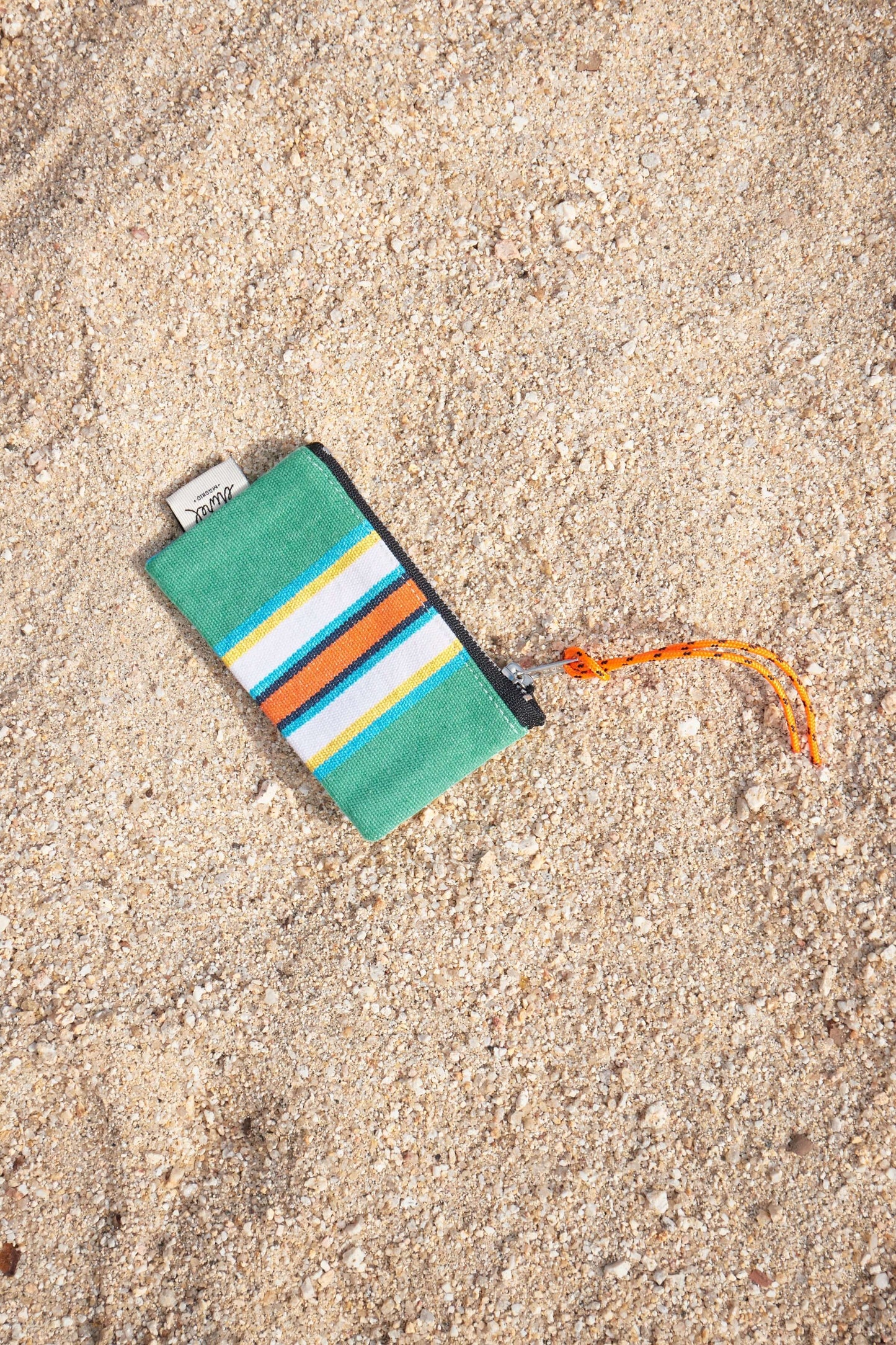 Monedero de lona verde a rayas con cremallera y cordón naranja, apoyado sobre arena de playa.