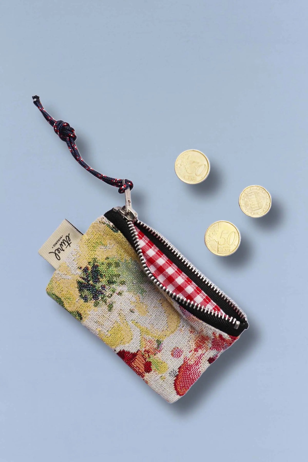 Small jacquard coin purse with poppy print, red gingham lining, zipper closure, cord pull, and three floating coins.