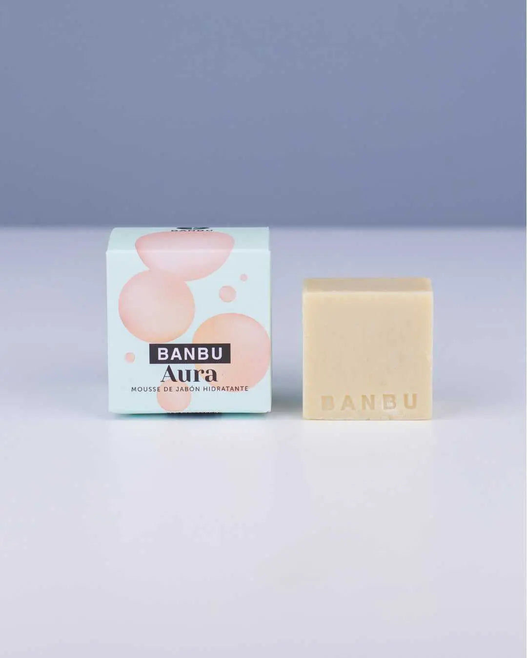 Mousse facial sólida Banbu Aura, junto a su caja de cartón decorada con burbujas rosas sobre fondo verde claro.